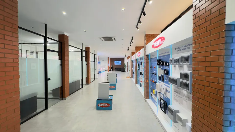 Allée centrale du showroom : murs d'exposition TOSUNLUX éclairés en bleu à droite, bureaux vitrés à gauche, coffrets présentés sur socles.